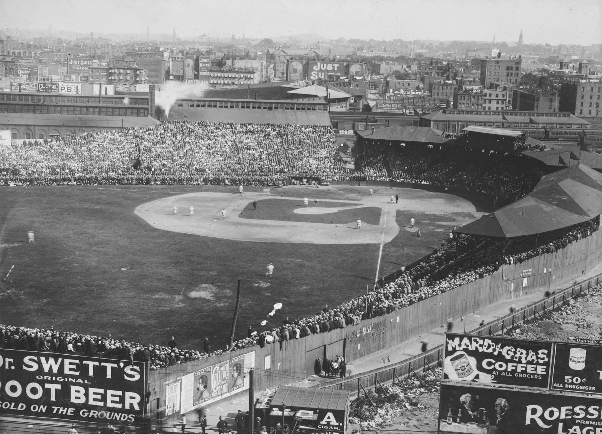 Black and white historical photo of a crowded early 20th-century baseball stadium, with packed stands, players on the field, and large vintage advertisements for products like Dr. Swett's Root Beer and Mardi Gras Coffee lining the outfield fence; city buildings with billboards are visible in the background.