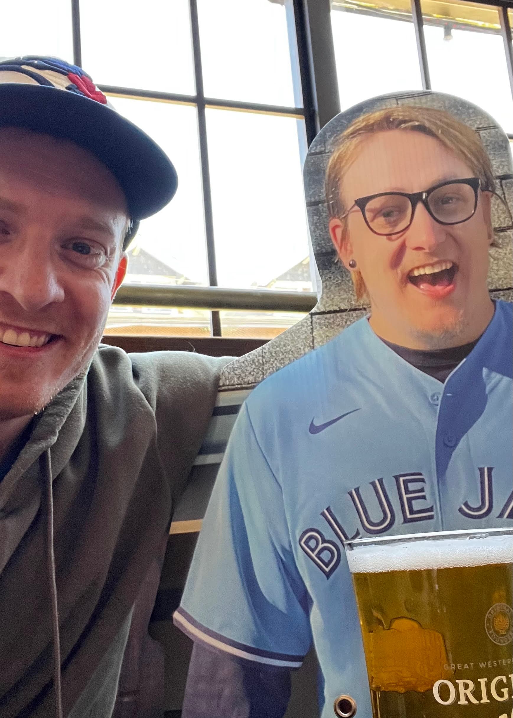 A smiling person (me) in a baseball cap and hoodie stands next to a life-size cardboard cutout of someone in a blue Toronto Blue Jays jersey and glasses (Iain). In the foreground, there is a glass of beer labeled Great Western Original. Large windows in the background let in natural light.